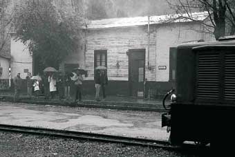 Tren y pasajeros en la estación, bajo la lluvia.